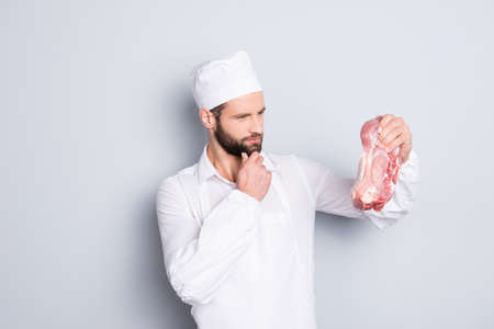 Portrait of pensive thoughtful butcher having fresh meat in hand looking at tenderloin holding hand on chin isolated on grey backgroundの写真素材