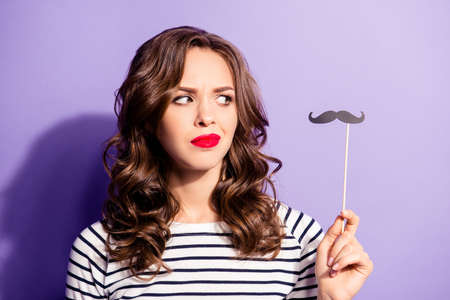 Portrait of unhappy confused girl with red lipstick looking at black carton cutout paper mustache on stick in hand isolated on violet backgroundの写真素材