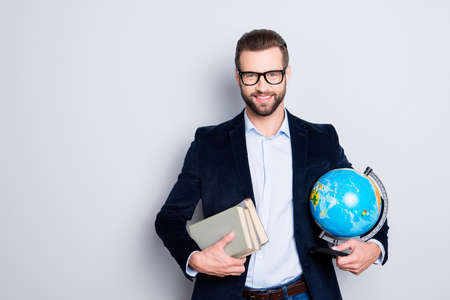 Portrait of stylish joyful teacher in shirt jacket with stubble having books and round globe, earth in hands, looking at camera, isolated on grey backgroundの写真素材