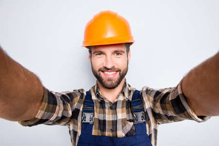 Portrait of handsome cheerful repairer in safety helmet with stubble shooting selfie on smart phone with two hands, wearing shirt and overalls, isolated on grey backgroundの写真素材