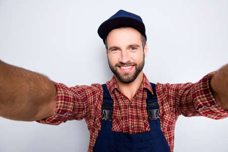Self portrait of joyful cheerful mechanic with stubble in blue overall, shirt shooting selfie on front camera with two hands, isolated on grey backgroundの写真素材