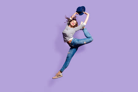 Portrait of active pretty girl in action jumping in the air showing perfect stretching making athletic ballet pose trick isolated on violet background holding hat in hands enjoying dreamdayの写真素材