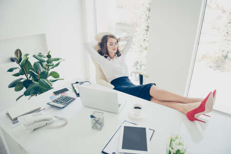 Pretty charming stylish trendy boss putting legs on the table holding hands behind the head with close eyes dreaming about vacation weekend having fun pleasure in modern office with interiorの写真素材