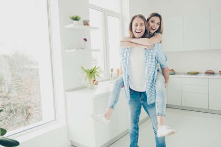 Portrait of funky trendy partners kidding in modern kitchen with interior, handsome man carrying on back lover looking at cameraの写真素材