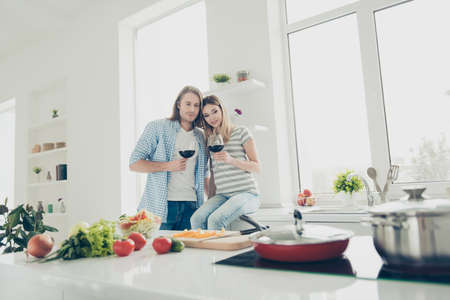 Portrait of dreamy romantic couple drinking alcohol beverage while preparing lunch in modern white kitchen celebrating 8-march. Affection feelings delight harmony conceptの写真素材