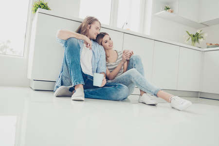 Low angle view of lovely romantic couple holding cups with coffee in hands enjoying weekend holiday together sitting on floor in modern white kitchen. Idyllic daydream harmony conceptの写真素材