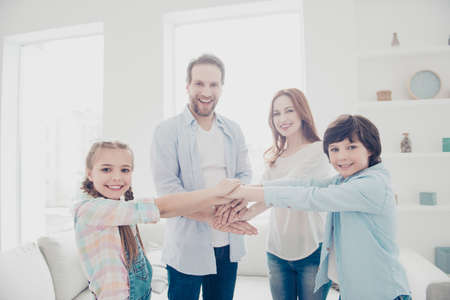 Portrait of cheerful joyful family with two kids standing in white living room putting hands on top...