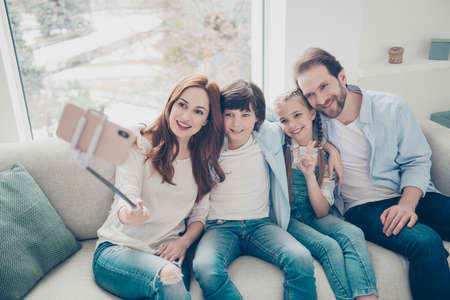 Portrait of friendly bloggers mom dad two kids sitting on sofa using smart phone gadget monopod shooting selfie in front camera gesturing v-sign peace symbol with two fingers having video-callの写真素材