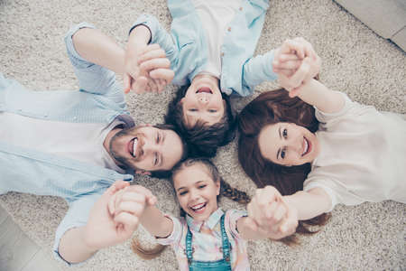 Top view portrait of cheerful positive comic funny family with two kids having good mood atmosphere holding hands up lying on carpet head to head. Trust support understanding conceptの写真素材