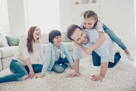 Portrait of daddy standing on knees carrying on back daughter mom and son sitting on the floor laughing enjoying game. Free-time daydream domestic lifestyle upbringing care conceptの写真素材