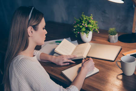Portrait of concentrated busy girl using book writing down notes in notepad with pencil.