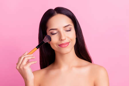Sensual charming pretty woman applying blusher on her cheekbones using make-up brush with close eyes isolated over pink backgroundの写真素材