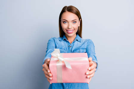 Portrait of cute charming girl presenting a gift in pink packaging with white bow standing over grey...