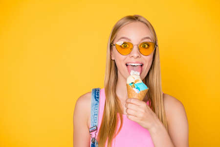 Portrait of impressed big-eyed girl in eyeglasses licking vanilla caramel ice cream scoops with tongue out looking at camera isolated on yellow backgroundの写真素材
