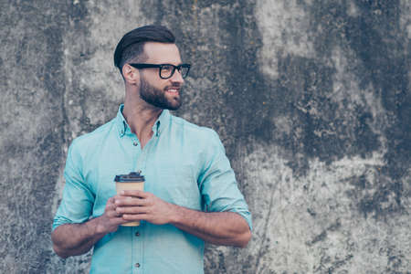 Side view portrait with copyspace of funky macho with modern hairdo having carton cup with coffee in hands looking away isolated on grey stone background. Barista caffeine cafe conceptの写真素材