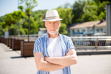 Portrait of concentrated dreamy businessman in straw hat holding arms crossed looking at camera standing over blurred street backgroundの写真素材