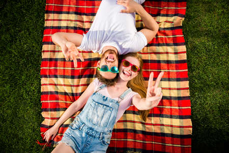 Top view portrait of cheerful funky couple lying on green grass head to head showing v-sign with two...