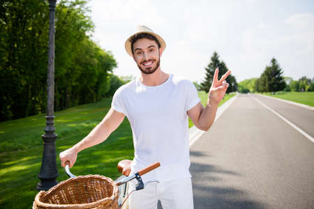 Portrait of cheerful positive attractive man in white outfit having bike gesturing two fingers v-sign with hand looking at camera. Harmony daydream serenity conceptの写真素材