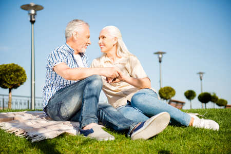 Portrait of sweet cute grandma and granddad in casual outfits sitting on plaid outdoor holding hands looking at each other enjoying weekend togetherの写真素材