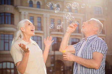 Portrait of funny playful spuses spending free time together, handsome man blowing soap bubbles for excited amazed lover over blurred building background outdoorの写真素材