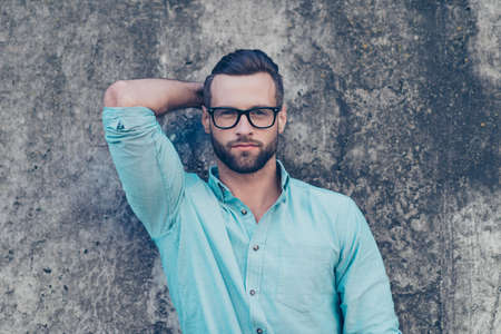 Portrait of stunning perfect guy with modern hairstyle in classic blue clothes holding hand behind head looking at camera isolated over grey stone wall. Model fashion beauty conceptの写真素材