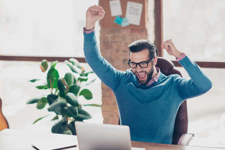 Stunning, smart, lucky, yelling man with raised arms, celebrating successfully completed work, project, presentation, sitting on armchair at desk in work stationの写真素材