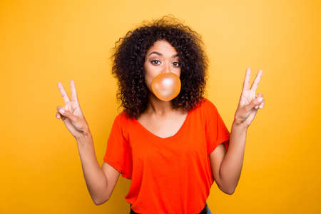 Portrait of cheerful cool girl with modern hairdo blowing chewing bubble gum gesturing v-signs with two hands looking at camera isolated on yellow backgroundの写真素材