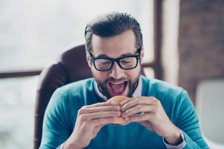 Close up portrait of cheerful busy excited handsome hungry manager doing to bite yummy appetizing cheeseburger in his hands sitting on armchair at workstationの写真素材