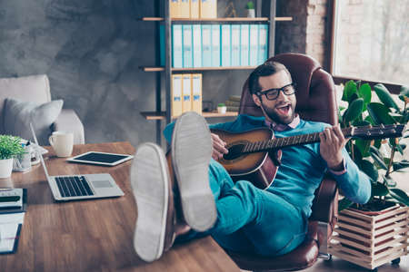 Cheerful, creative man sitting at desktop on chair, putting legs on table, having guitar in hands,...