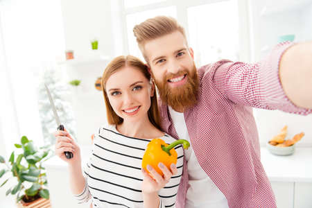 Healthy nutrition veggie cook blogger video-call concept. Self portrait of joyful partners shooting selfie on front camera holding knife and fresh yellow pepper in hands having online meetingの写真素材