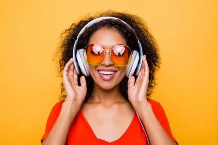 Head shot portrait of cheerful positive girl in summer glasses with headphones on head listening favorite pop music isolated on yellow backgroundの写真素材