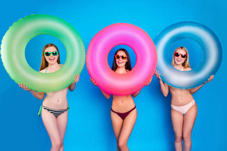 River sea water sunbathing suntan tan concept. Portrait of funny playful girls holding colorful swim rings in hands having thin slim legs isolated on vivid blue backgroundの写真素材