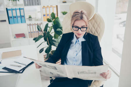 Portrait of clever attractive woman in eyeglasses reading newspaper about policy sitting in modern...