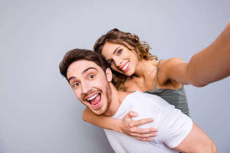 Rest relax leisure concept. Self portrait of positive cheerful couple, excited man carrying on back pretty lover shooting selfie on front camera isolated on grey backgroundの写真素材