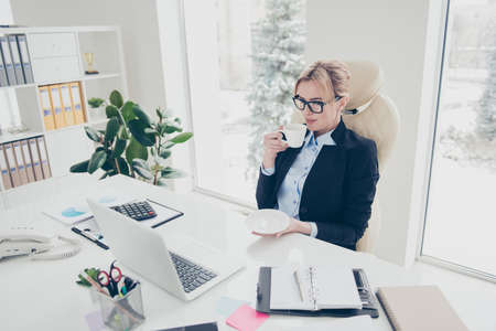 Portrait of stylish pretty lawyer in shirt jacket drinking hot beverage enjoying free time watching movie video using laptop sitting in modern white workstation with interiorの写真素材