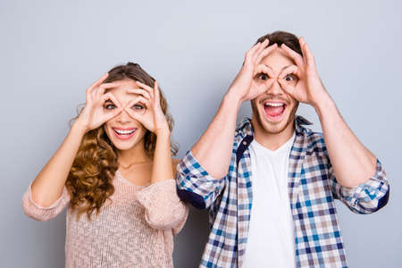 Portrait of childish cool couple making binoculars with fingers around eyes keeping mouth open isolated on grey background. Entertainment conceptの写真素材