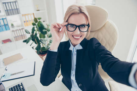 Portrait of stylish trendy woman in eyeglasses sitting in modern workstation shooting selfie on front camera enjoying break having online meeting wearing classy jacket shortの写真素材