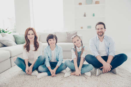 Portrait of cheerful cute four people sitting in modern white apartments on the floor near couch with crossed legs looking at camera wearing jeans sneakers casual outfitsの写真素材
