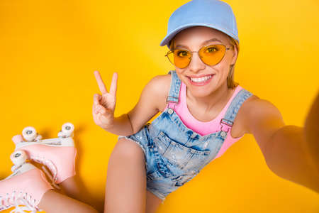 Self portrait of positive toothy girl shooting selfie on front camera wearing roller skates gesturing v-sign with hand isolated on yellow background, fitness workout athletic sport conceptの写真素材