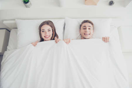 Portrait of young attractive sleepy couple awakening together in bed in the morning, covered with blanket, girl and guy smiling with their teeth. Top above high angle viewの写真素材