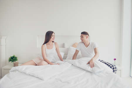 Portrait of young attractive cute couple wearing pajama, sitting together in bed, smiling, looking...