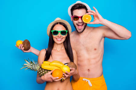 Handsome couple in in a good mood and colourful eyewear holding a lot of fruit. Funky young woman and handsome men messing around with fruitの写真素材