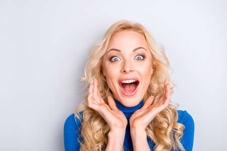 Portrait of sexy, surprised, impressed, stylish, pretty, cute, charming, nice, blonde woman in blue turtleneck  with wide open eyes and mouth looking at camera holding hands near faceの写真素材