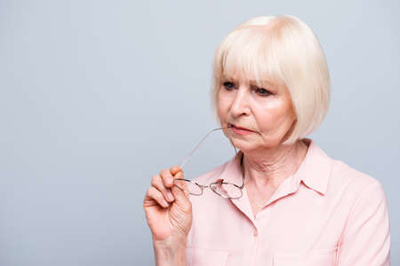 Portrait of old adult blonde caucasian sad depressed lady, holding eye glasses, thinking over grey background, isolated. Copy spaceの写真素材
