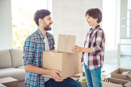 Young happy smiling family two persons daddy and son wearing casual holding carton boxes with stuff things in light studio living room, moving to new flatの写真素材