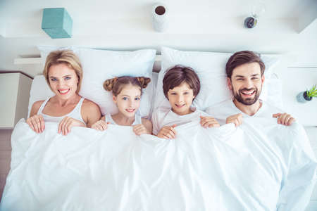 Young happy smiling family four persons lying together in bed laughing covered with blanket...