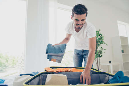 Time to fly for the holidays! Happy and joyful young man packing jeans in a suitcaseの写真素材