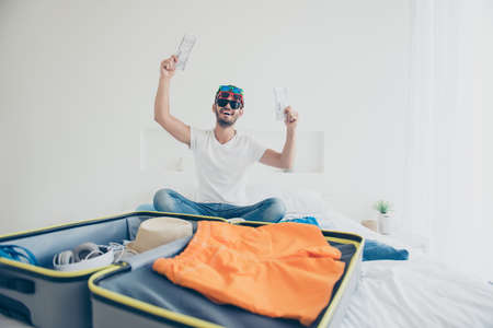 Full-legh full-body portrait of happy young man holds in his hands hot tickets for the plane sitting...