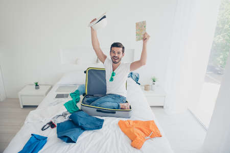 Full-legh full-body portrait of happy guy put the suitcase on the bed in the bedroom and sit into it waiting for a trip to the resortの写真素材