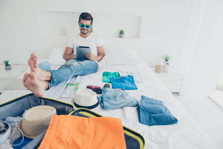 Time of holidays! Full-legh full-body portrait of happy young man looks at the tablet laughs and...